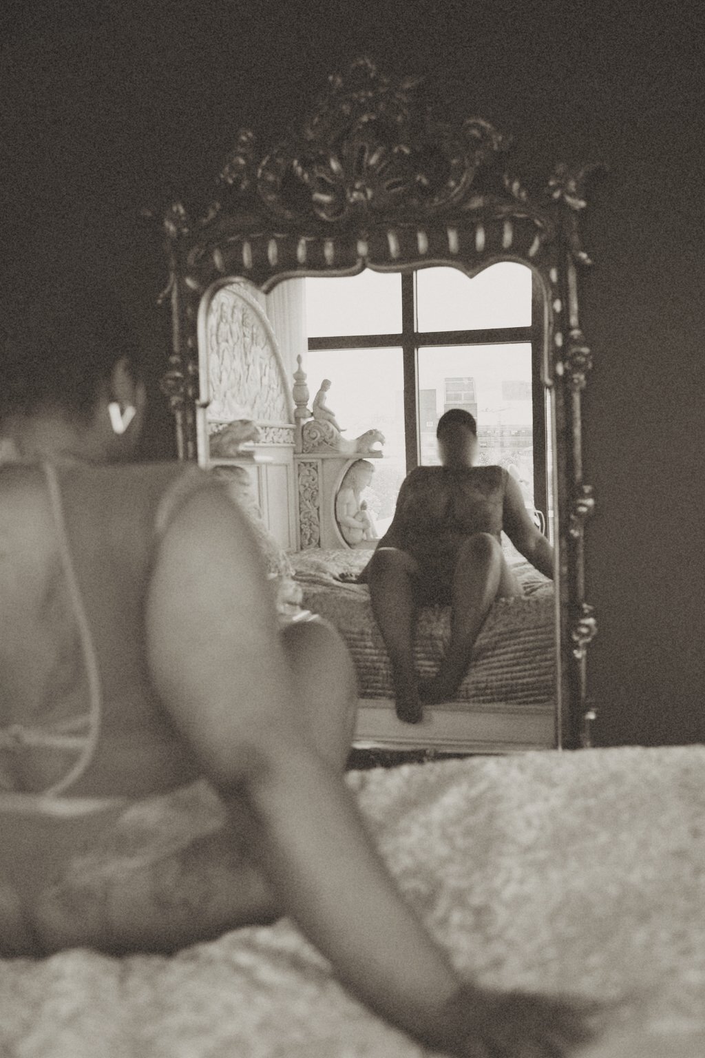 Black and white image of Sable Gold, Kinky GFE in lace body, sitting on a bed looking into a large mirror furry hoodie, holding riding crop in front of mirror, commanding dominatrix Luxembourg